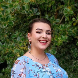 Millie-Jo faces the camera, smiling. She has her hair pinned up and wears a floral blue dress, with greenery behind her.